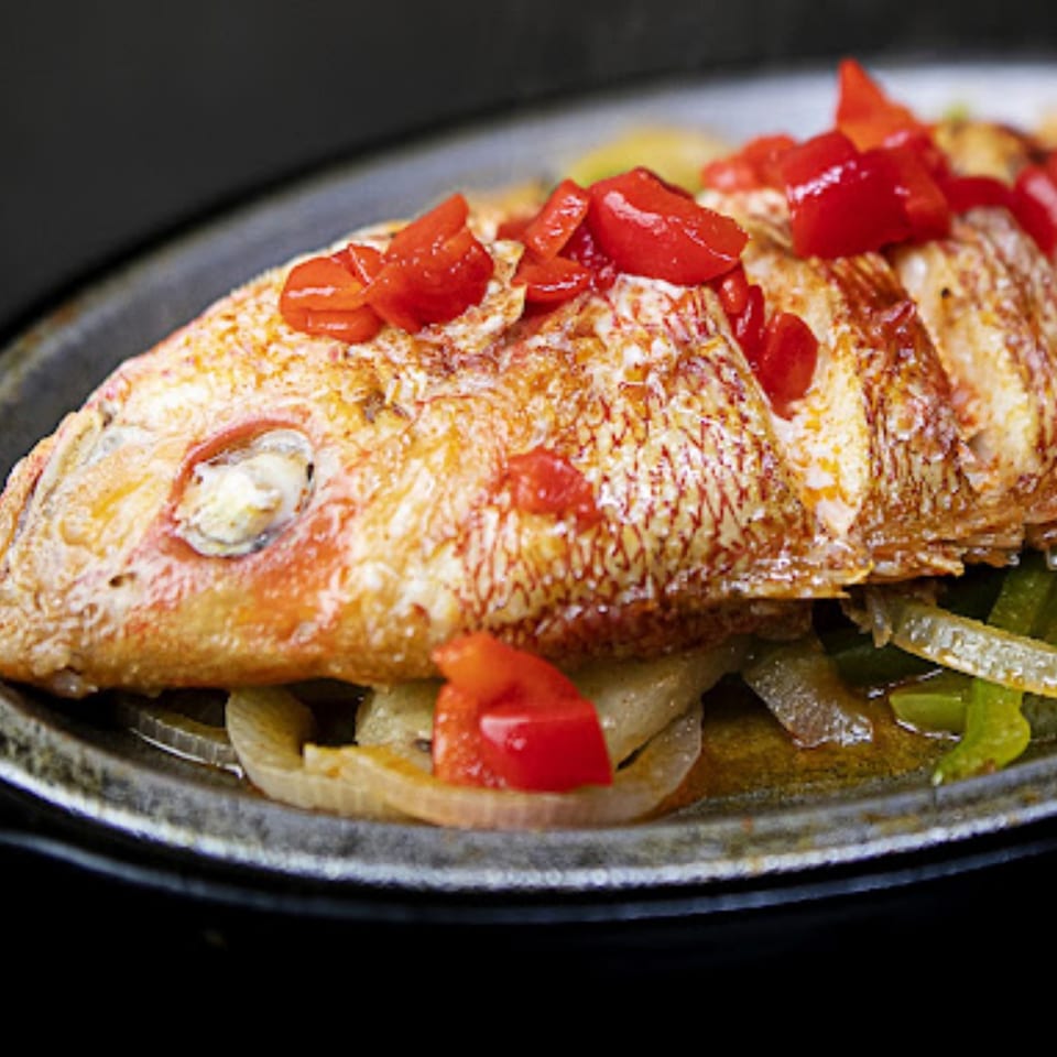 Whole fried fish topped with red bell peppers and onions, served on a dark plate.
